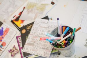 A cup with crayons and markers in it, on a table with scattered papers and drawings from children